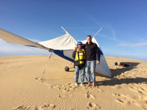 Two people pose for a picture in front of a hang glider on the dunes at the Outer Banks